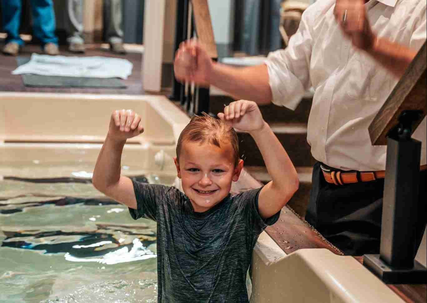 A boy raises both arms with a grin as he comes up out of the baptismal water, the pastor beside him