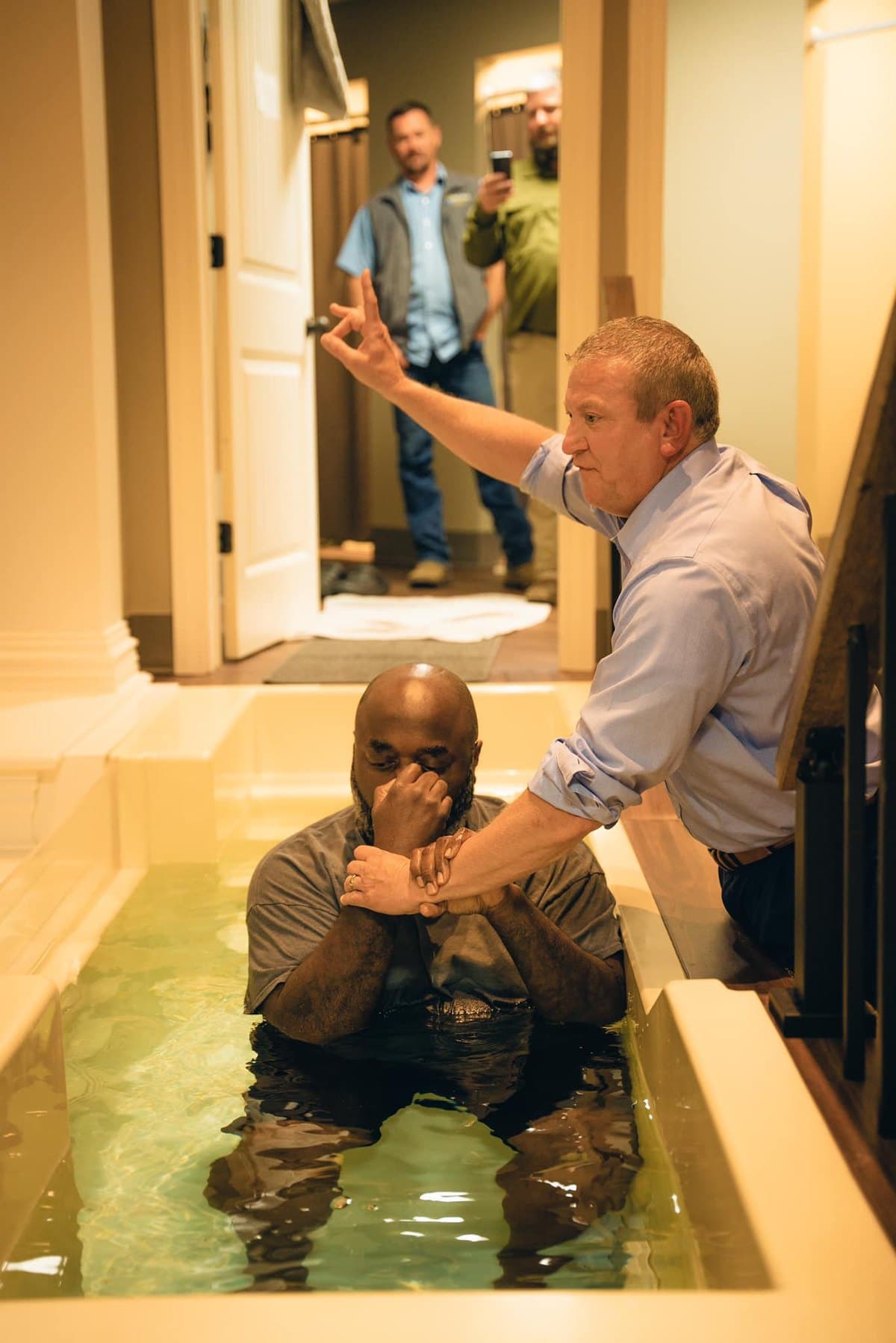 A man being baptized, standing in the water with the pastor's hand raised beside him