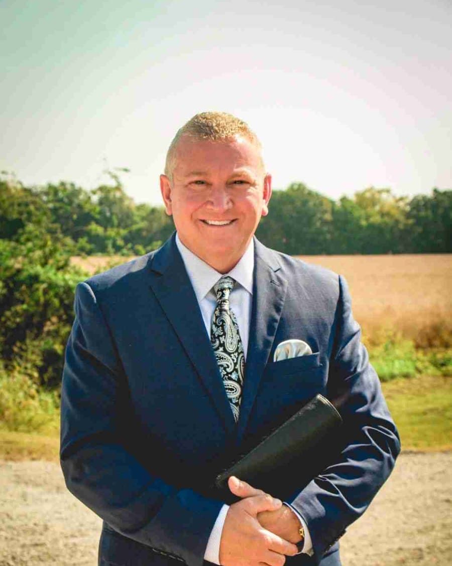 Bro. Curt Howard, pastor of Maple Springs Missionary Baptist Church, standing at the edge of a field in a navy suit with his Bible under his arm